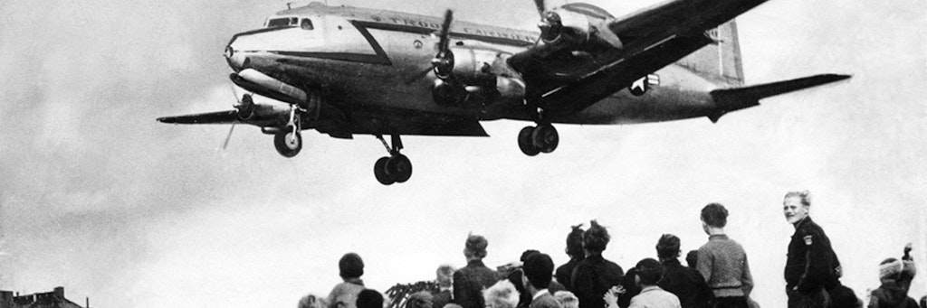 Ein Rosinenbomber im Anflug auf den Flughafen Tempelhof – über wartende Kinder. Links im Bild das Luftbrückenhaus.