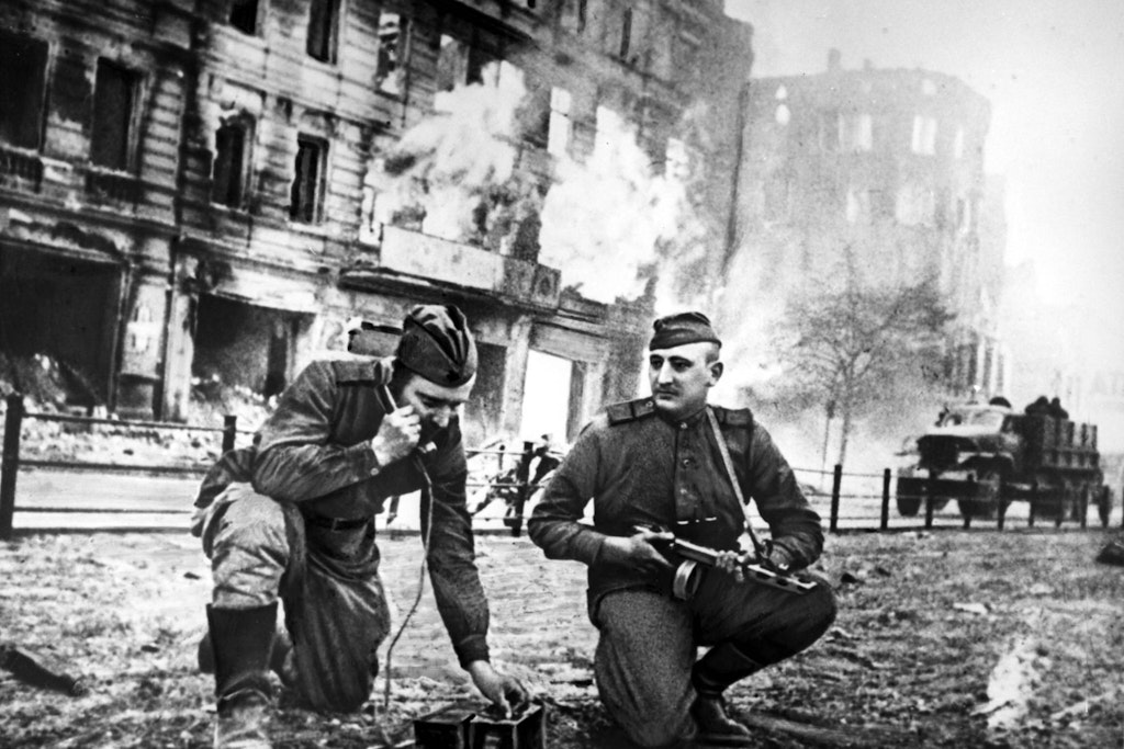 Zwei sowjetische Soldaten machen in einer Berliner Straße Meldung. Im Hintergrund steht ein Haus in Flammen. Eine gestellte Szene?