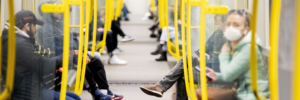 Fahrgäste sitzen in einer U-Bahn, wobei eine Frau einen Mundschutz trägt. 