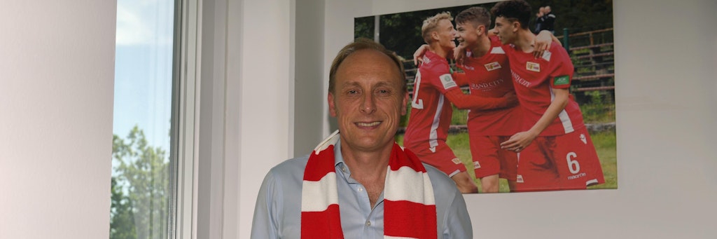 Andrew Wallis in seinem Büro. Im Hintergrund ein Bild von Unions A-Junioren. Dort war er schon engagiert, ehe Aroundtown nach dem Aufstieg bei den Eisernen als Haupt- und Brustsponsor einstieg. 