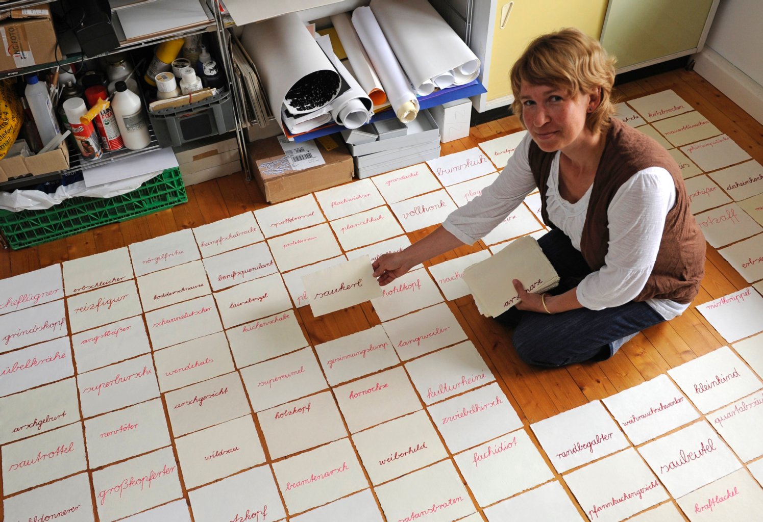 Die Künstlerin Ingke Günther kniet in Gießen in ihrem Atelier auf dem Boden, um ein Blatt Büttenpapier mit einem aufgestickten Schimpfwort auf den Boden zu legen.Ob neumodische Klassiker wie «Kotzbrocken» oder der «Poussierstängel» aus Omas Zeiten: Schimpfwörter faszinieren Günther. Die Künstlerin hat eine Sammlung mit inzwischen fast 1500 Wörtern.