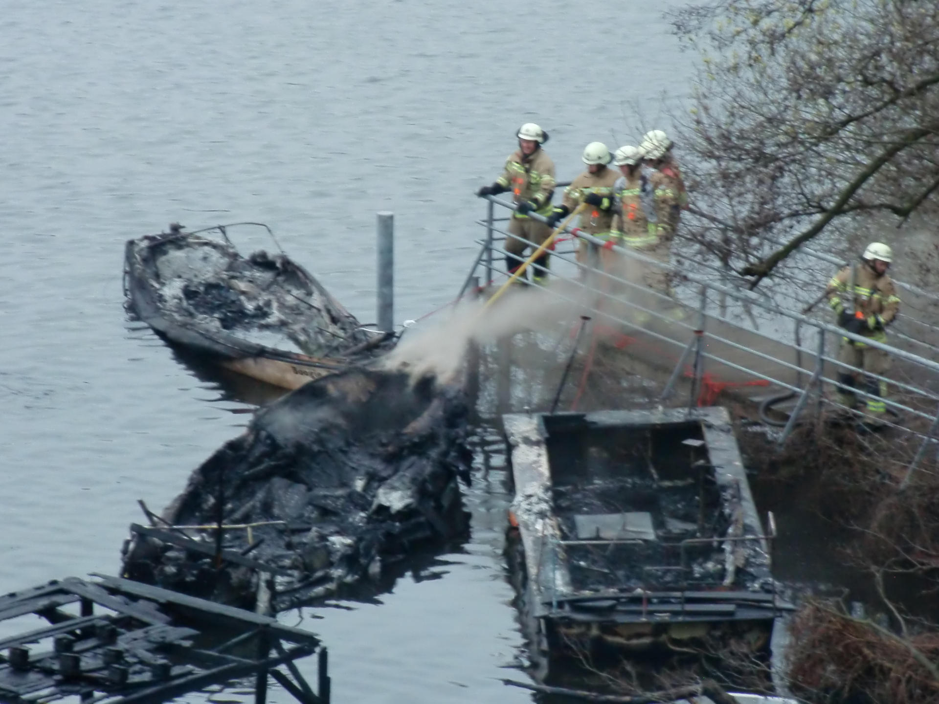 Feuerwehrleute löschen ein bereits völlig verkohltes Boot vom Ufer aus.
