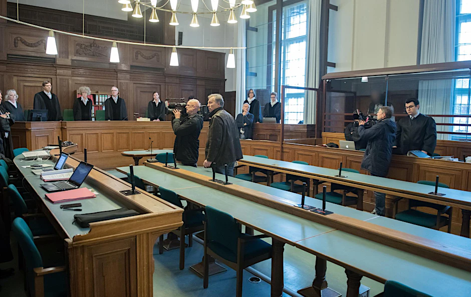 Ein Angeklagter (hinten, 3.v.r.) steht in einem Gerichtssaal hinter einer Glasscheibe. Mit weiteren drei Männern muss er sich wegen mutmaßlicher Unterstützung des IS verantworten.