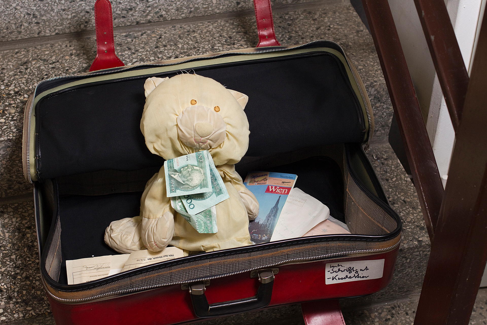 Ein selbstgenähter Teddy sitzt in einem Koffer, hat zwei 1000-Forint-Scheine auf der Brust. Das Schmusetier war 1989 als Schmuggler unterwegs. Eine Familie aus der DDR, im Urlaub in Ungarn, hatte die schon bestehende Möglichkeit genutzt, nach Wien zu reisen und wieder zurückzukehren. Das Geld, das sie nicht hätten ausführen dürfen, war im Teddy versteckt.  