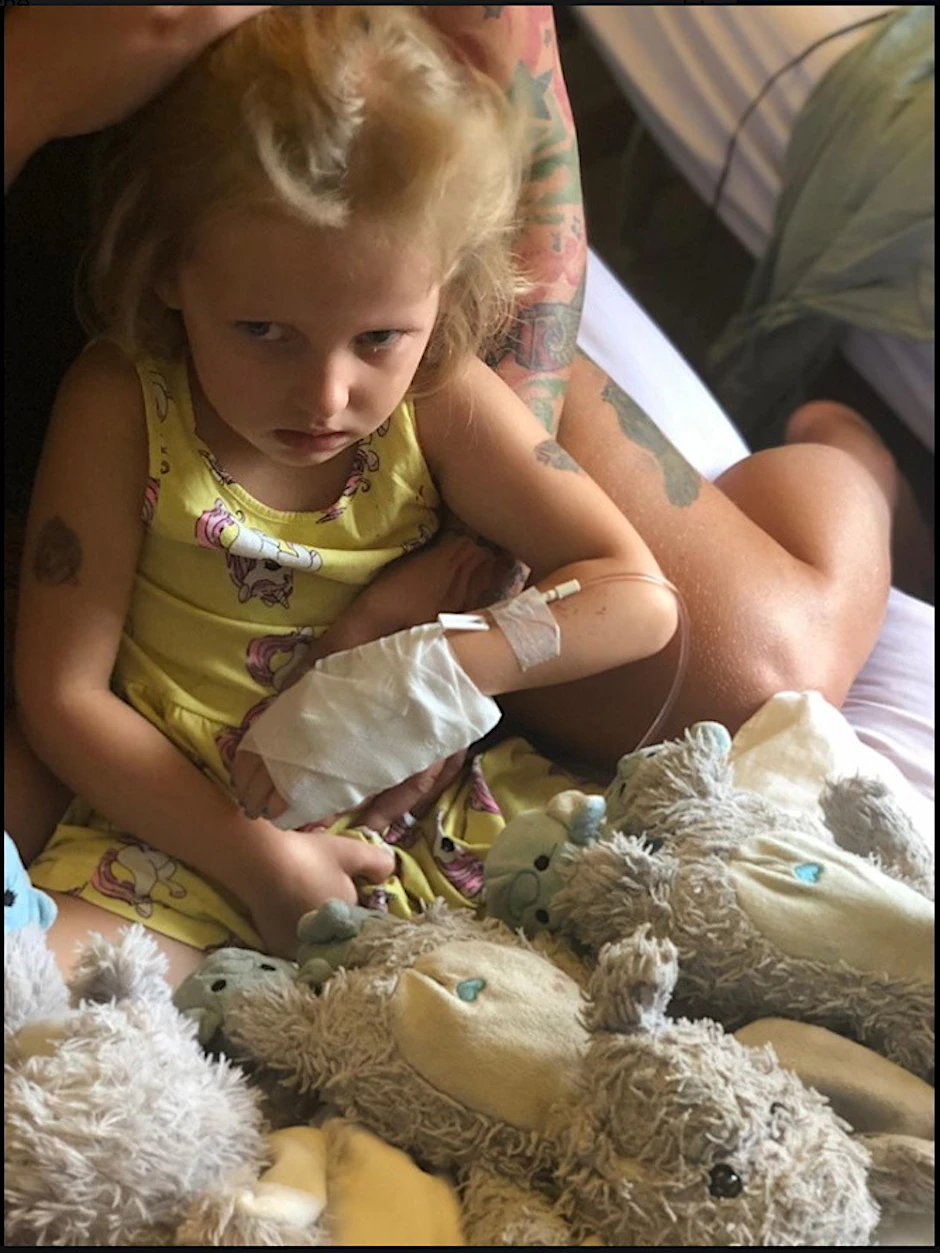 Die kleine Lotta guckt trotzig. Sie trägt einen Verband an der Hand und hängt am Tropf.