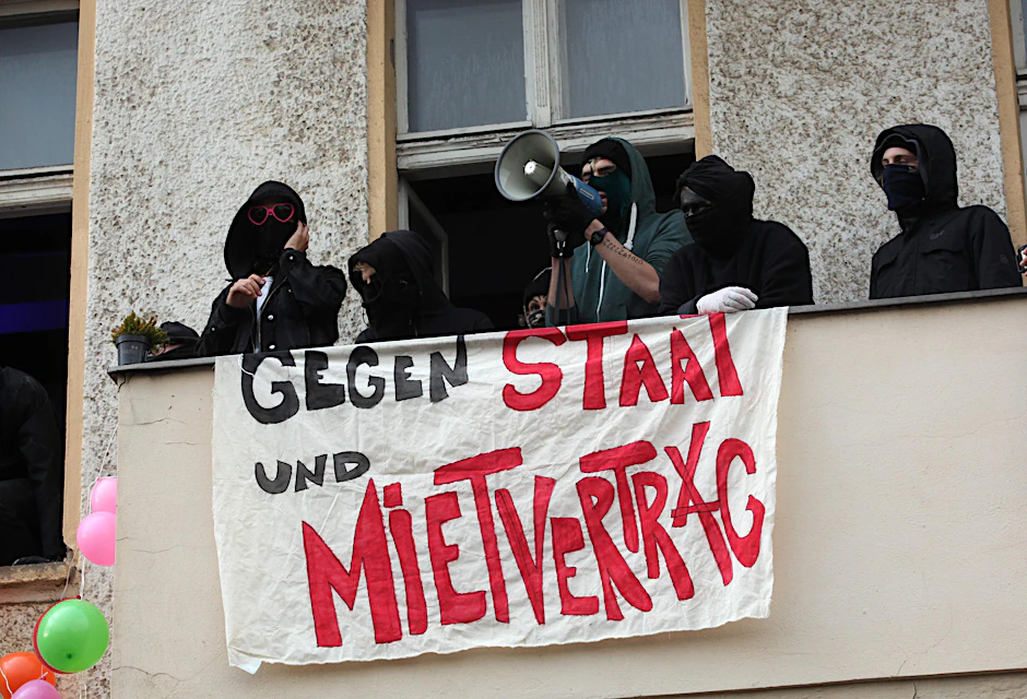 Maskierte Besetzer haben in der Frankfurter Allee ein Transparent aufgehängt, ein Mann spricht durchs Megafon zu den Demonstranten vor dem besetzten Haus.