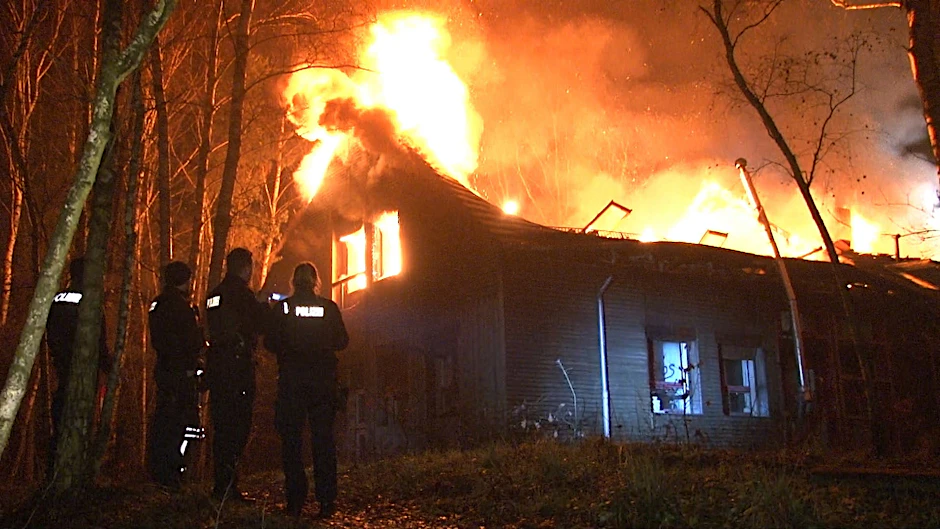 Einsatzkräfte der Polizei stehen vor dem lichterloh brennenden Gebäude. 