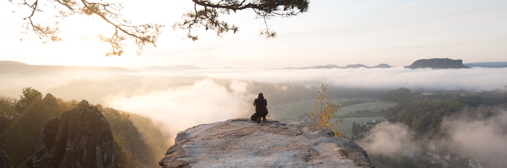 Ein Mann hockt im Nationalpark Sächsische Schweiz auf dem Basteifelsen und fotografiert den Sonnenaufgang.