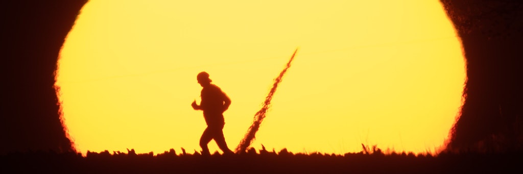 Eine Frau joggt am frühen Morgen beim Sonnenaufgang über einen Feldweg im Landkreis Hildesheim, während vor der Sonne ein Rotorblatt eines Windrades zu erkennen ist.
