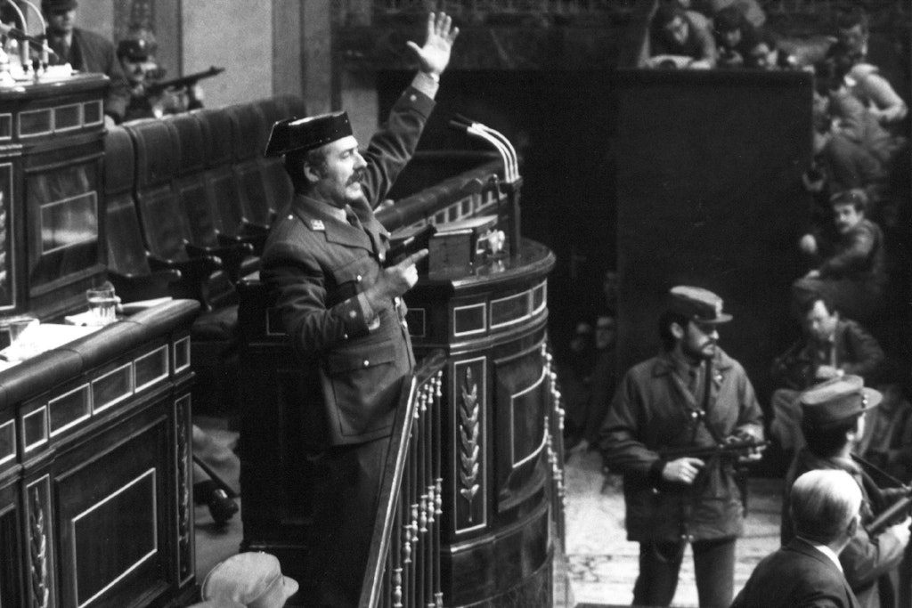 Putschversuch 1981: Mit der Pistole in der Hand steht ein Oberstleutnant der Guardia Civil auf dem Rednerpult des Parlaments, davor Uniformierte mit Maschinenpistolen. Fast alle Parlamentarier gingen in Deckung, nur Ministerpräsident Adolfo Suarez und zwei Abgeordnete blieben stehen oder sitzen und stellten sich so den Putschisten entgegen.