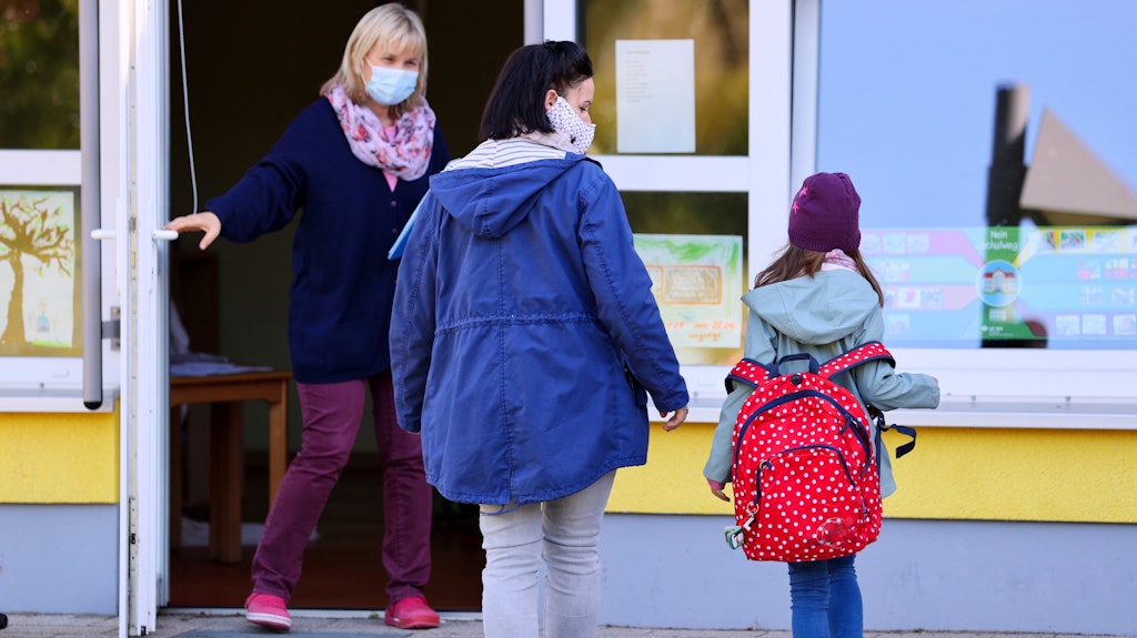 Eine Mutter bringt ihre Tochter zur Tür einer Kindertagesstätte, wo sie bereits von einer Erzieherin erwartet wird. Denn Eltern dürfen wegen der Corona-Schutzmaßnahmen die Kita nicht betreten. 