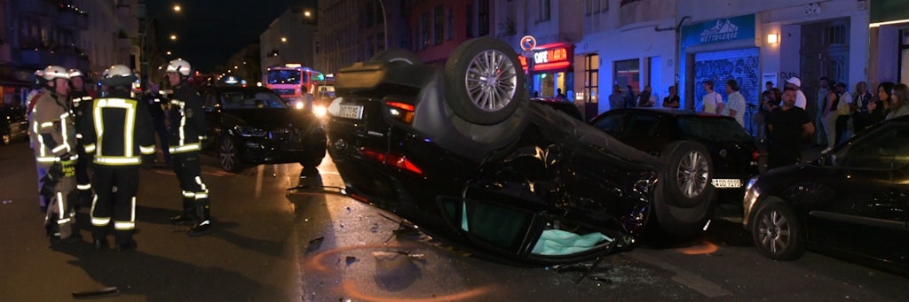 Ein Auto liegt mitten in Neukölln auf dem Dach. Ein andere Fahrzeug hatte den Hyndai gerammt. Links stehen die Retter, rechts Schaulustige. <br><br>