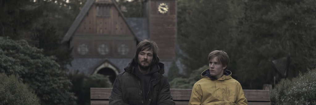 Szene aus „Dark“: Die Schauspieler Andreas Pietschmann und Louis Hofmann sitzen vor der Holzkirche auf dem Stahnsdorfer Friedhof.