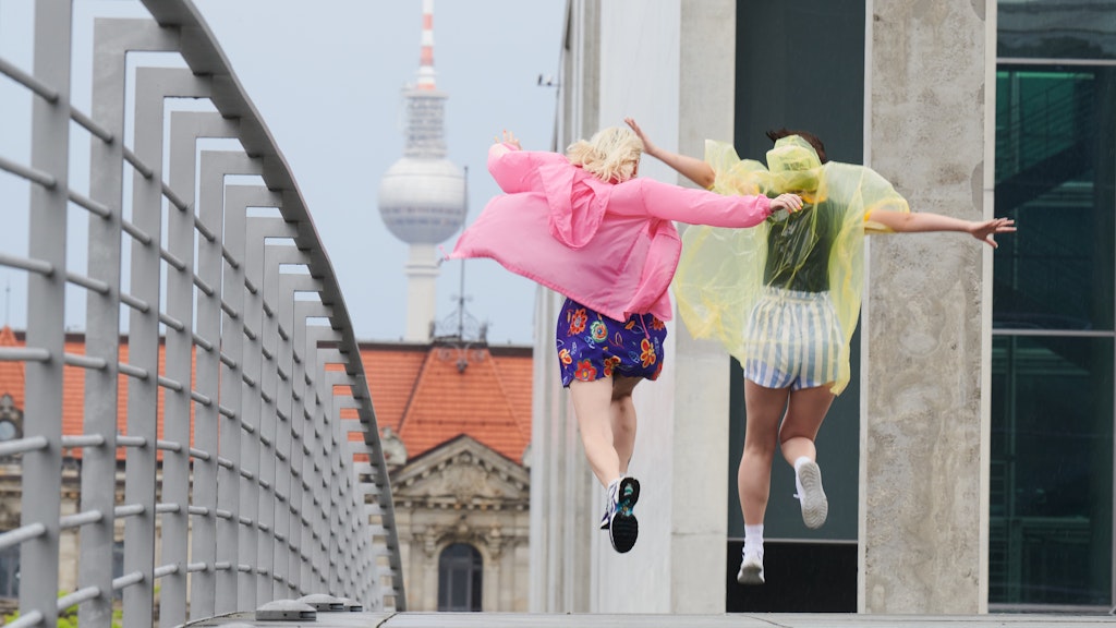 Zwei junge Frauen springen nach einem Regenguss mit bunten Regencapes über den Marie-Elisabeth-Steg am Paul-Lübbe Haus. 