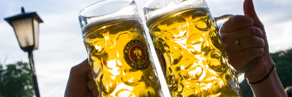 Zwei Personen prosten sich in einem Biergarten mit zwei Maßkrügen zu. 