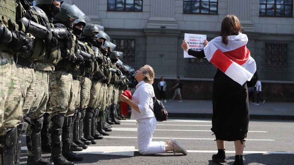 Eine Frau kniet in Minsk vor einer Reihe Männer in Tarnanzügen, eine zweite hat eine alte belarussische Fahne umgelegt, die als Symbol der Opposition gilt.
