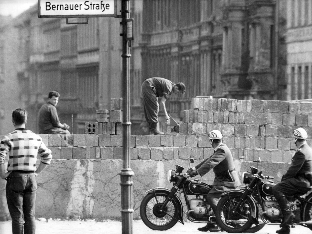 Mauerbau 1961: Stein für Stein wächst die Mauer, die Ost-Berliner Arbeiter im August des Jahres an der Bernauer Straße errichten. West-Berliner Polizisten und Passanten sehen dem ungläubig zu.