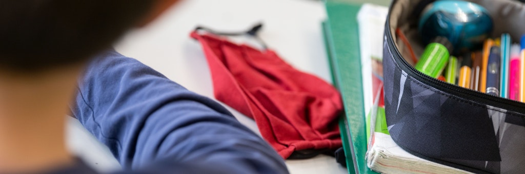 Eine Mund-Nasen-Bedeckung liegt während einer Unterrichtsstunde neben einem Mäppchen und Schulbüchern auf einem Schultisch.