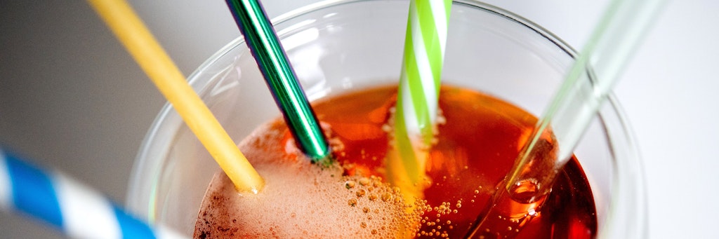 Ein Glas Limonade mit mehreren wiederverwendbaren Trinkhalmen aus Papier, Edelstahl, Hartplastik und Glas sowie einer als Trinkhalm verwendeten Nudel (2. v. l.) steht auf einem Tisch. 