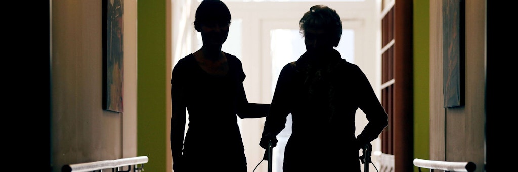 Eine Pflegekraft begleitet die Bewohnerin eines Altenheims mit Rollator beim Gang durch den Flur.