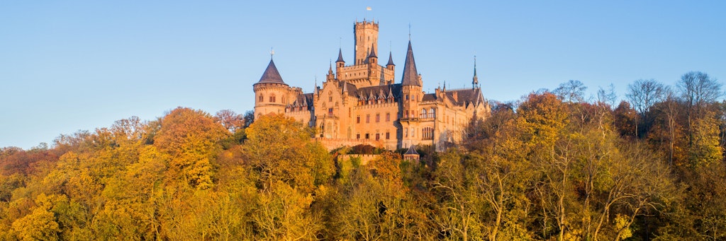 Das Schloss Marienburg in der Region Hannover steht im warmen Licht der aufgehenden Sonne inmitten herbstlich verfärbter Bäume.