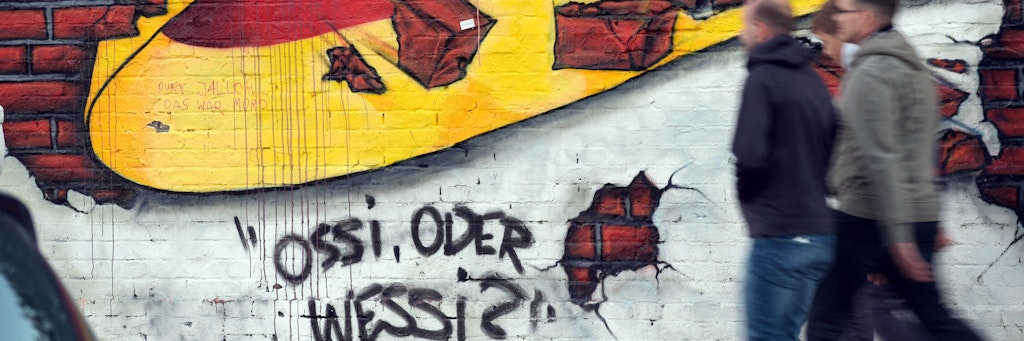 Berlin: Passanten gehen an einem Wandbild von Caspar Kirchner mit der deutschen Nationalflagge und dem Schriftzug "Ossi oder Wessi? vorbei. 