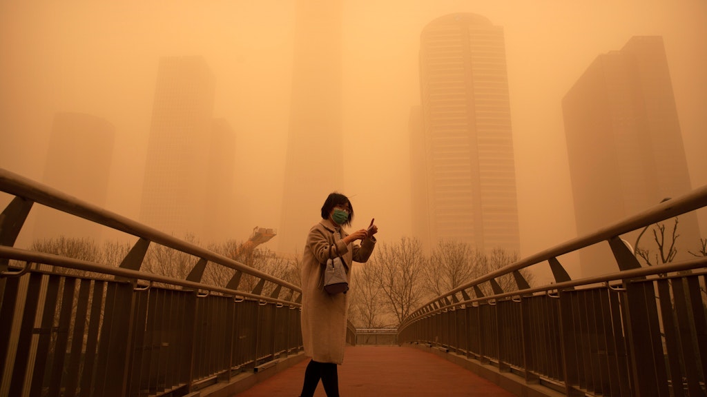 Eine Frau steht in Peking auf einer Brücke. Der Sandsturm hat die ganze Stadt in ein dunkles Orange gehüllt.