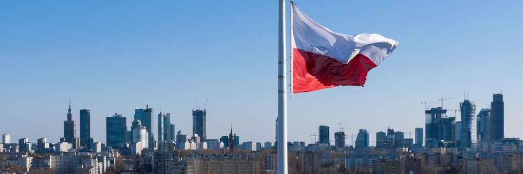 Eine polnische Nationalflagge weht auf einer wenig befahrenen Straße im Wind vor der Warschauer Skyline.