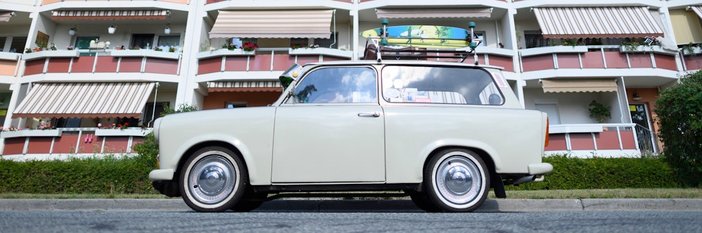 Ein Trabant Kombi 601s, der auf seinem Dachgepäckträger einen Koffer und ein Longboard geschnallt hat, steht vor einem Neubaublock.