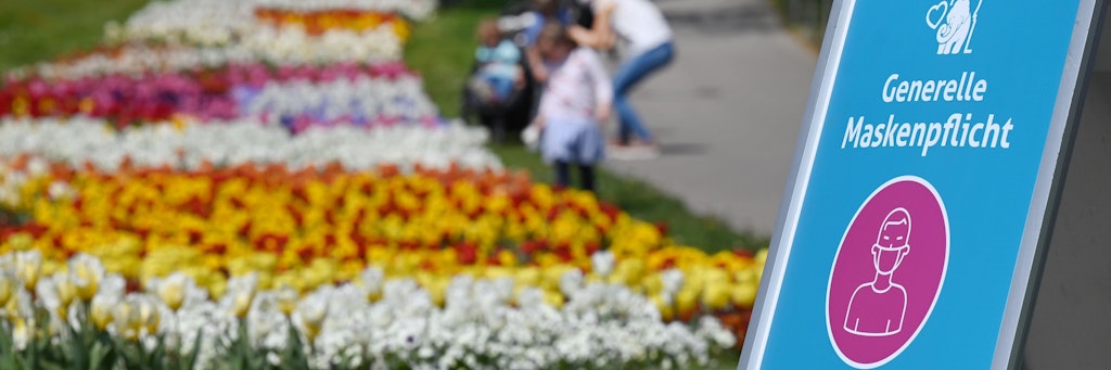 Spaziergänger gehen an einem farbenprächtigen Tulpenbeet vorbei, im Vordergrund steht dabei ein Hinweisschild mit der Aufschrift: „Generelle Maskenpflicht“.