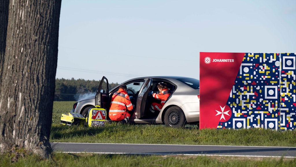Neben einer gestellten Unfallszene der Johanniter-Unfall-Hilfe ist eine Stellwand mit einem QR-Code zu sehen, der die Botschaft „Gaffen tötet“ enthält.