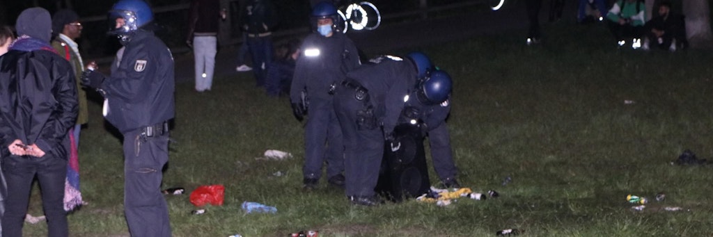 Polizisten heben in der Hasenheide eine Musikanlage hoch, umgeben von Flaschen und anderem Müll.