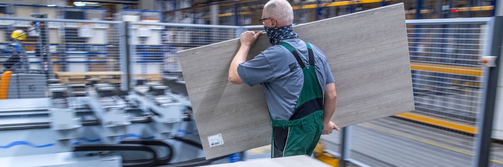 In einer Büromöbel-Fabrik schleppt ein Mitarbeiter eine Platte zur Bearbeitung mit Bohrer und Fräse.