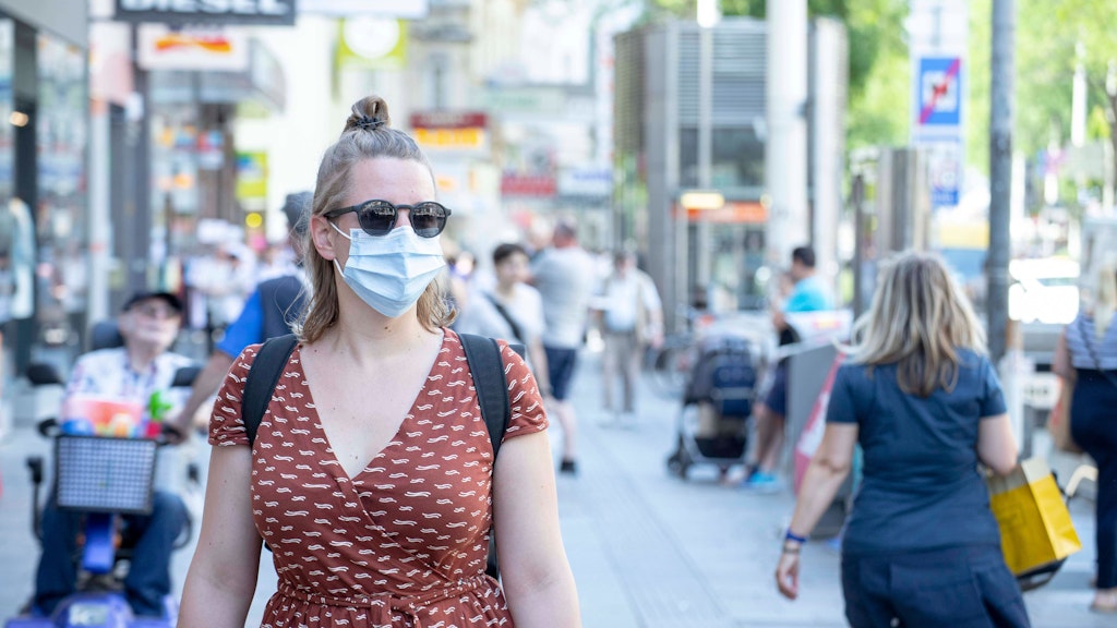 Eine junge Frau mit Sonnenbrille trägt einen blauen Mund-Nasen-Schutz in einer Fußgängerzone.