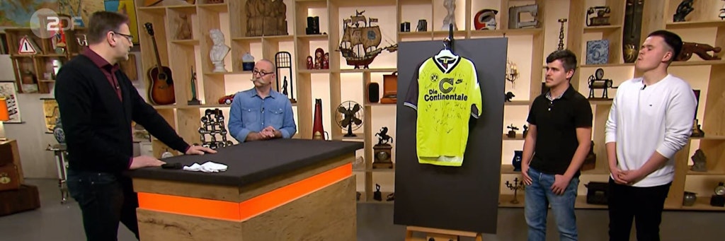 „Bares für Rares“-Experte Detlev Kümmel erklärt den Formalla-Brüdern und Horst Lichter, was es mit diesem Trikot auf sich hat.