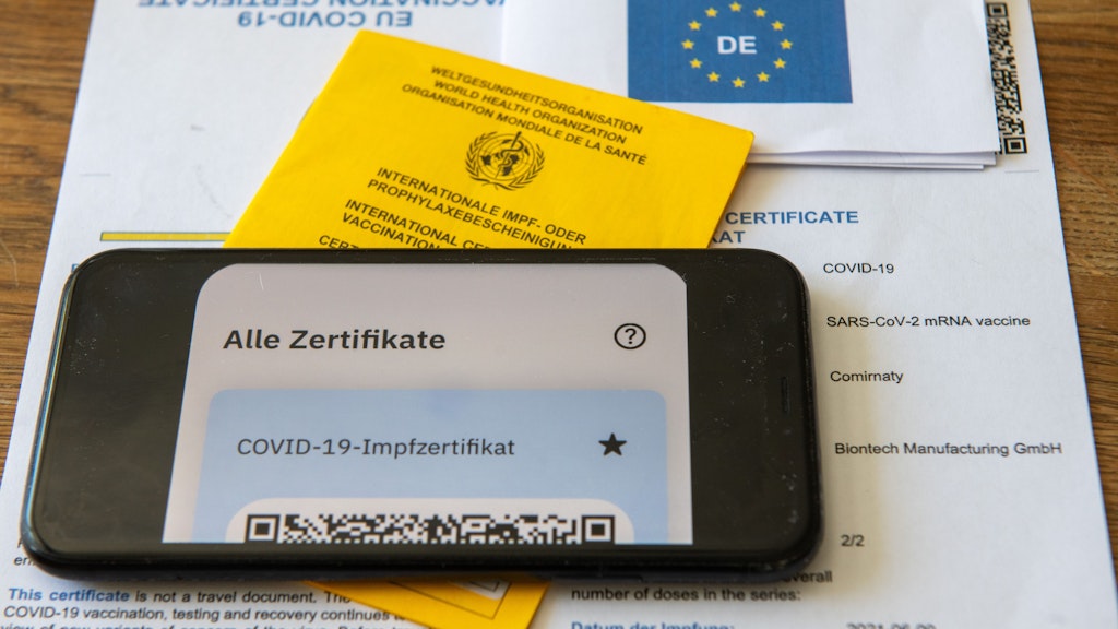 Ein Impfpass und ein Smartphone, auf dem die App CovPass läuft, liegen auf einem Impfzertifikat, das von einer Apotheke ausgestellt wurde.