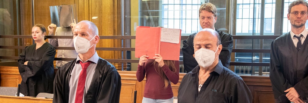 Zwei der Angeklagten stehen mit Aktendeckeln vor dem Gesicht hinter ihren Anwälten.