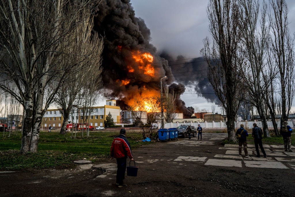 Personen stehen auf einer Straße und beobachten wie am Horizont Feuer und schwarzer Rauch aufsteigt, nachdem das russische Militär ein Treibstoffdepot beschossen hat.