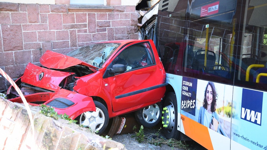 Ein zerstörtes Auto und ein Linienbus stehen nach einem Unfall an einer Hauswand.