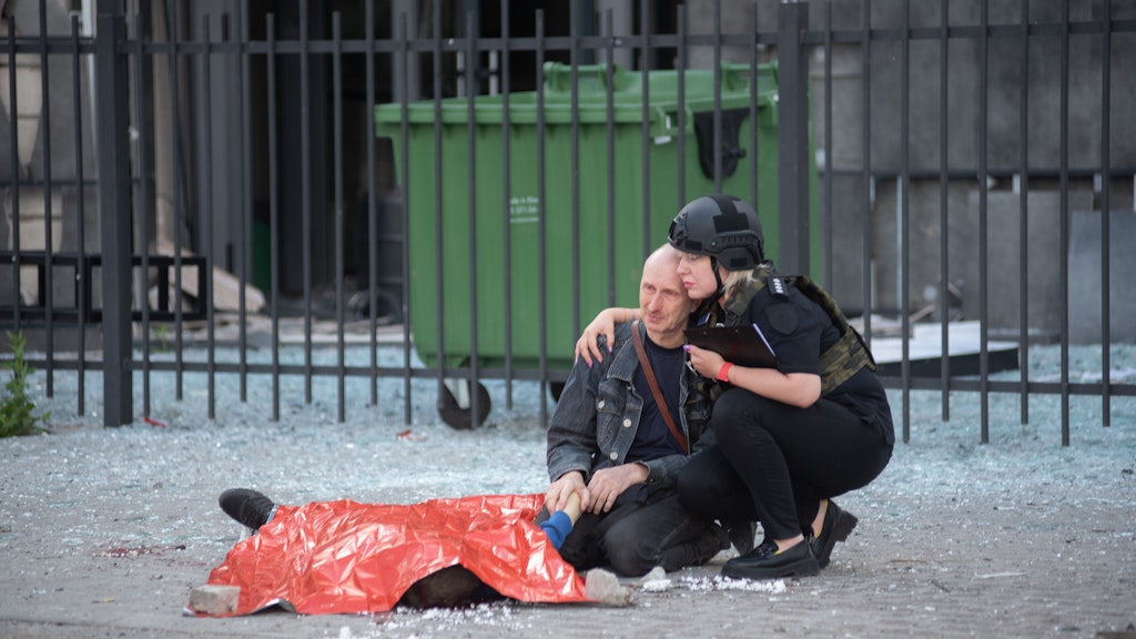 Eine ukrainische Polizistin hockt sich neben den Mann, der die Hand seines toten Sohnes hält.