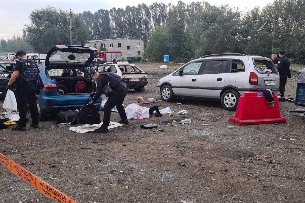 Eine Leiche liegt abgedeckt nach einem Raketenangriff auf dem Boden, während Polizisten an Autos Gepäck untersuchen. 