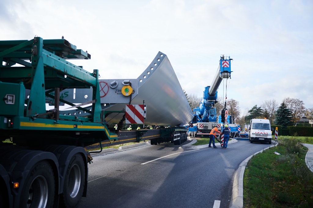 Ein Kran soll das Bauteil eines Windrads vom festgefahrenen Schwerlasttransporter heben.