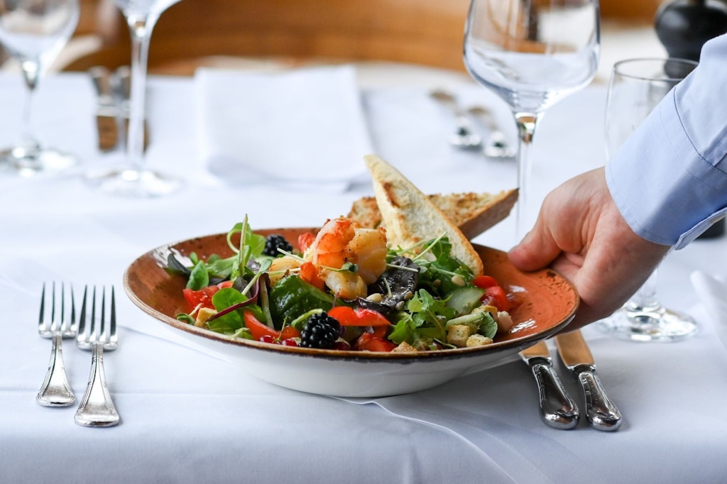  Ein Kellner serviert einen Salatteller in einem Restaurant. Ob er dafür ein Trinkgeld erhält, hängt davon ab, in welchem Land er serviert. 