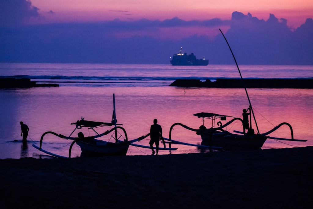 Männer befestigen am Strand von Nusa Dua auf Bali Boote, während sich der vor Sonnenaufgang mit schillernden Farben ankündigt.