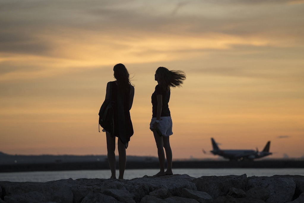 Zwei Frauen schauen dem Sonnenaufgang an einem Strand von Bali hinterher, im Hintergrund Flugzeuge.