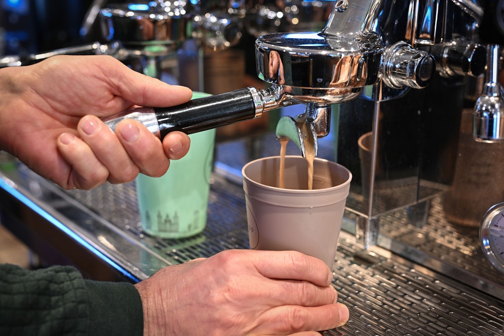 Ein Mitarbeiter eines Bio-Ladens bereitet einen Kaffee zu, der in einen Mehrwegbecher fließt.