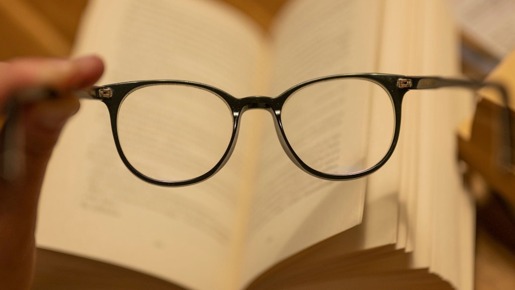 Ein Mann hält eine Lesebrille vor einem geöffneten Papierbuch. Die Abstände beim Arbeiten am Computer sind andere. 