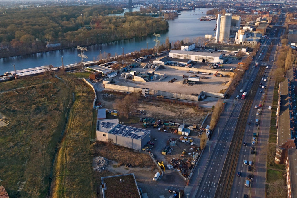Hier will die Firma Archigon ihr Spreewerk bauen: In etwa zwischen der vorderen vor dem Zementwerk querstehenden Hallen bis zur Tischlerei Maßwerk unten im Bild. Das leicht abgewinkelte Gebäude mit Solardach wird abgerissen, die ummauerte Baustoff-Recyclinganlage wird wegen heranschreitenden Rentenalters von den Eigentümern aufgegeben. Rechts an der Köpenicker Chaussee die Gaswerkssiedlung.