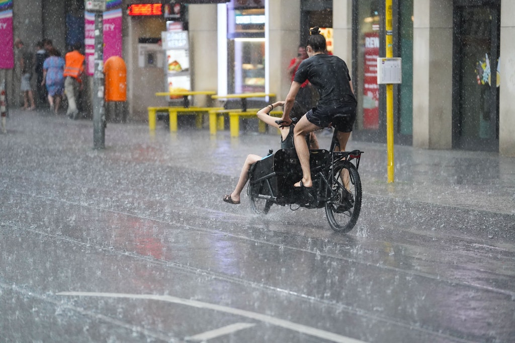 Junge Leute fahren mit einem Lastenfahrrad, während starker Regen auf die Friedrichstraße prasselt.