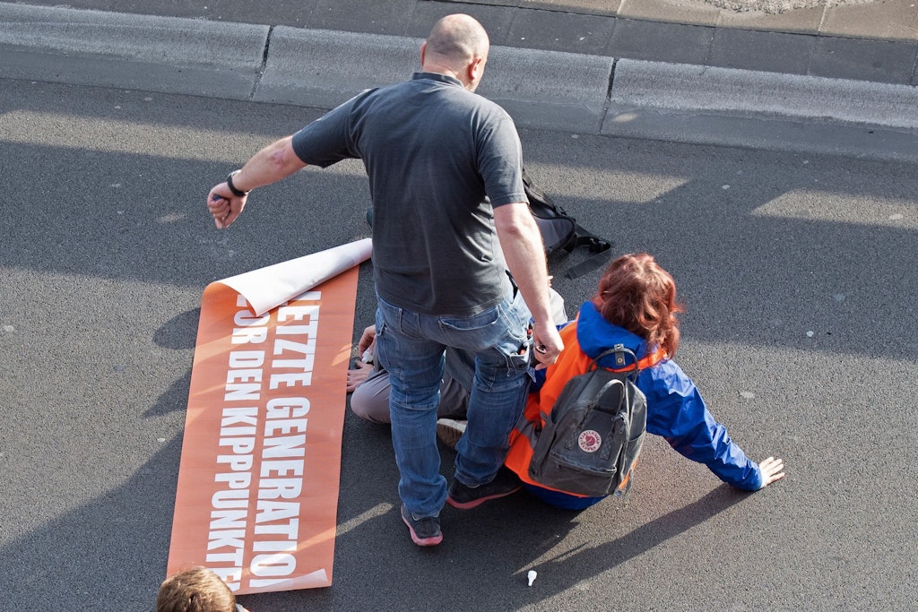 Ein wütender Autofahrer tritt eine Klima-Kleberin von der Straße.